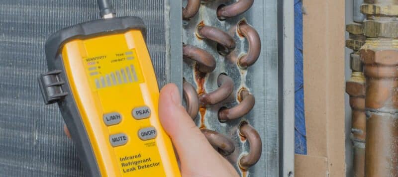 hand holding a yellow infrared refrigerant leak detector next to an ac unit evaporator coil